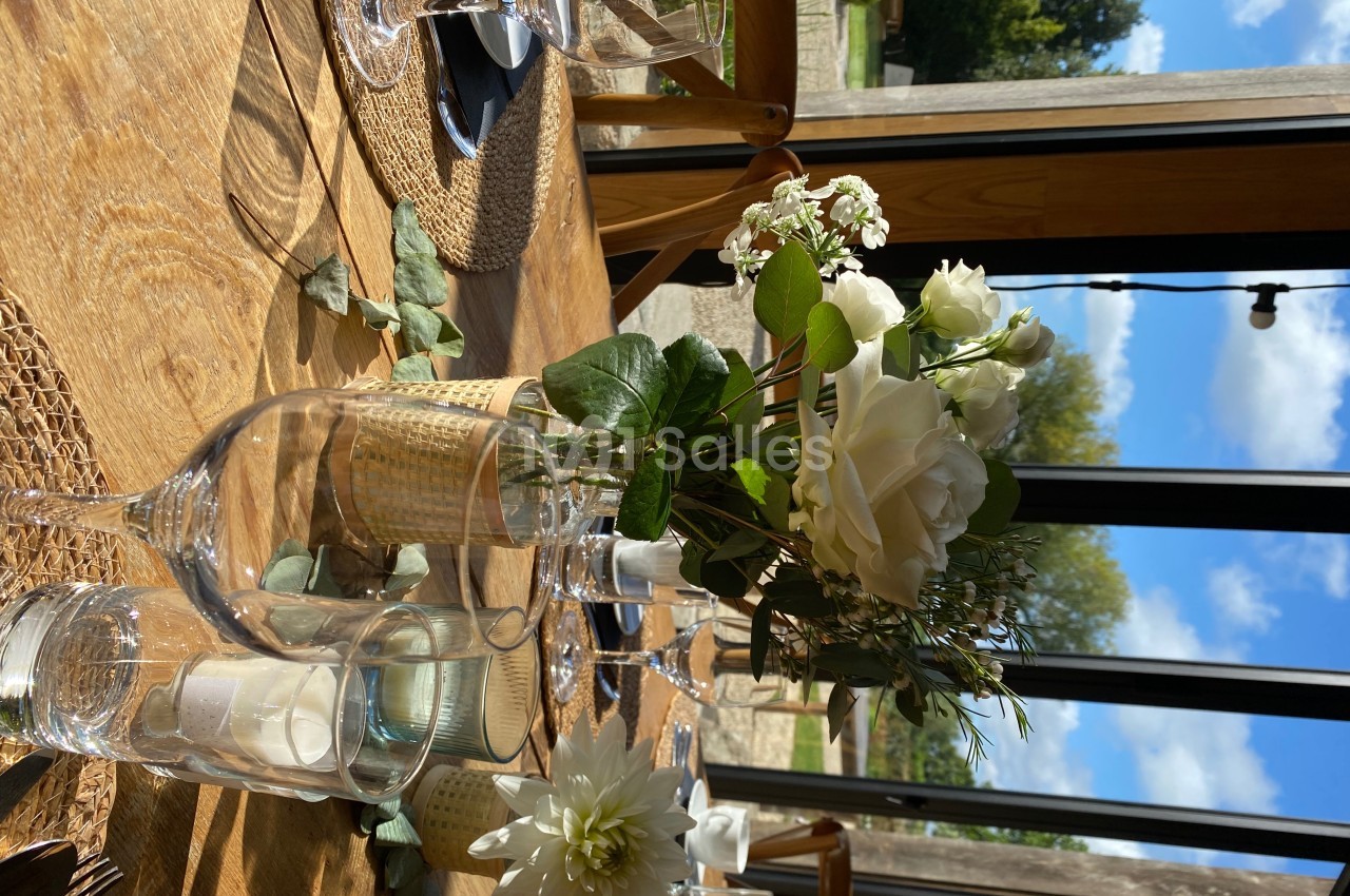 Table en bois dressée avec des verres, des bougies et un bouquet de fleurs blanches, près d'une grande baie vitrée.