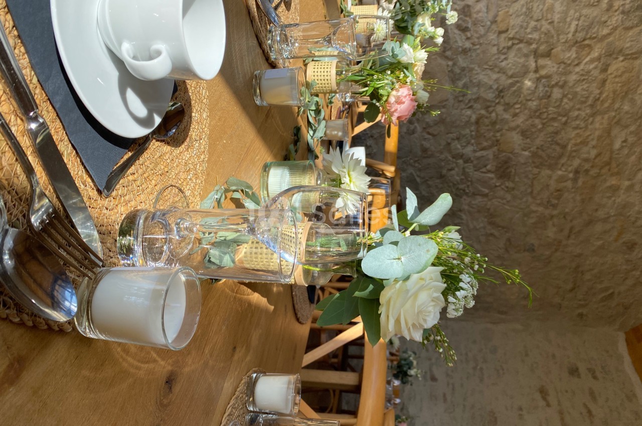 Table en bois dressée avec des verres, tasses blanches, bougies et décorations florales dans une salle en pierre.