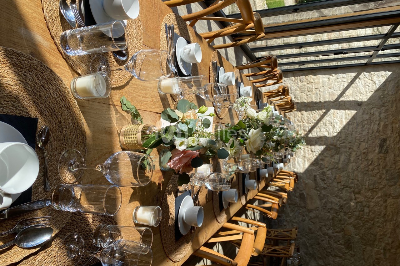 Table en bois dressée pour un repas, avec vaisselle élégante, verres, bougies et compositions florales au centre.
