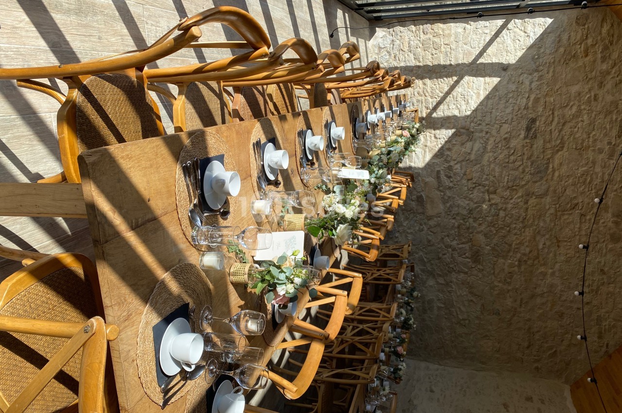 Table en bois dressée pour un repas, avec vaisselle blanche, décor floral et lumière naturelle entrant par une verrière.