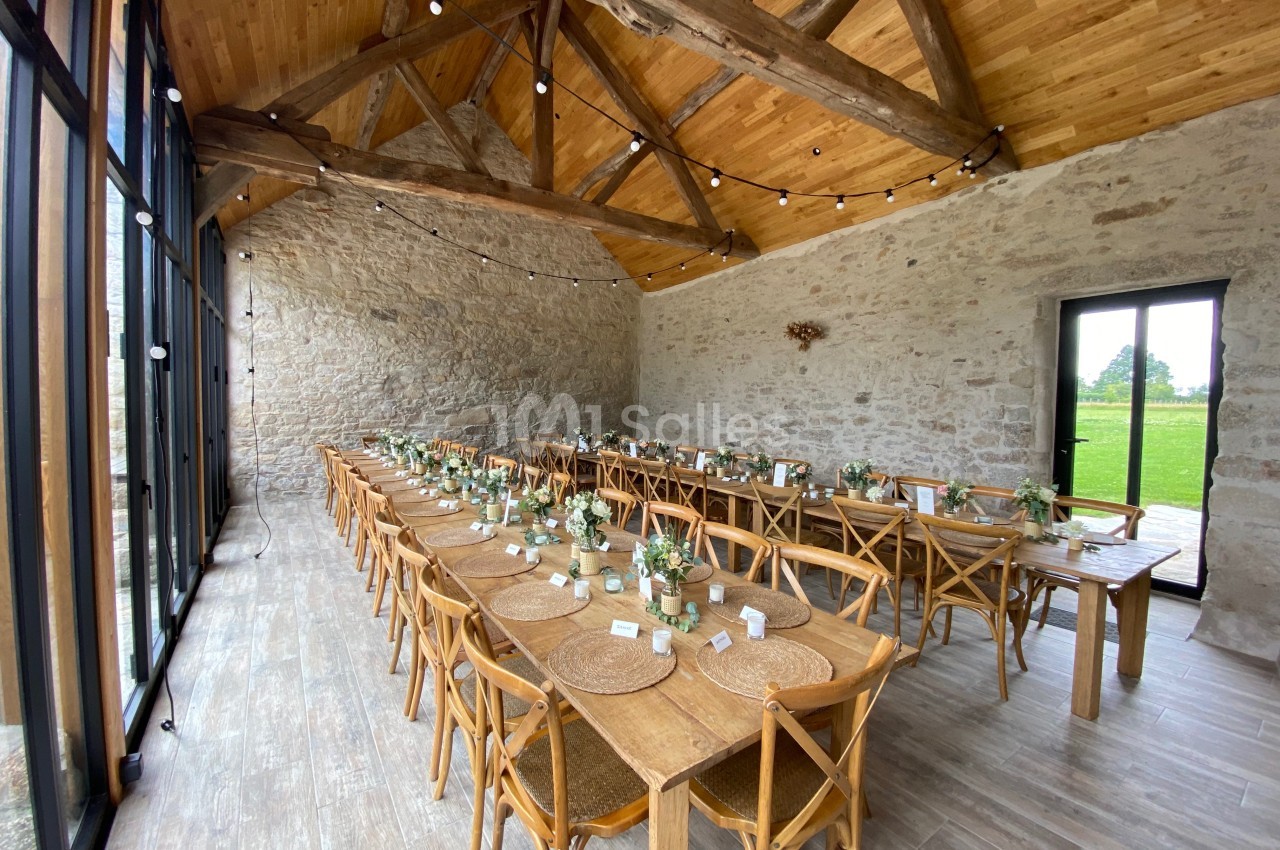 Salle en pierre avec poutres apparentes, tables en bois alignées et décorées de fleurs, éclairée par de grandes baies…