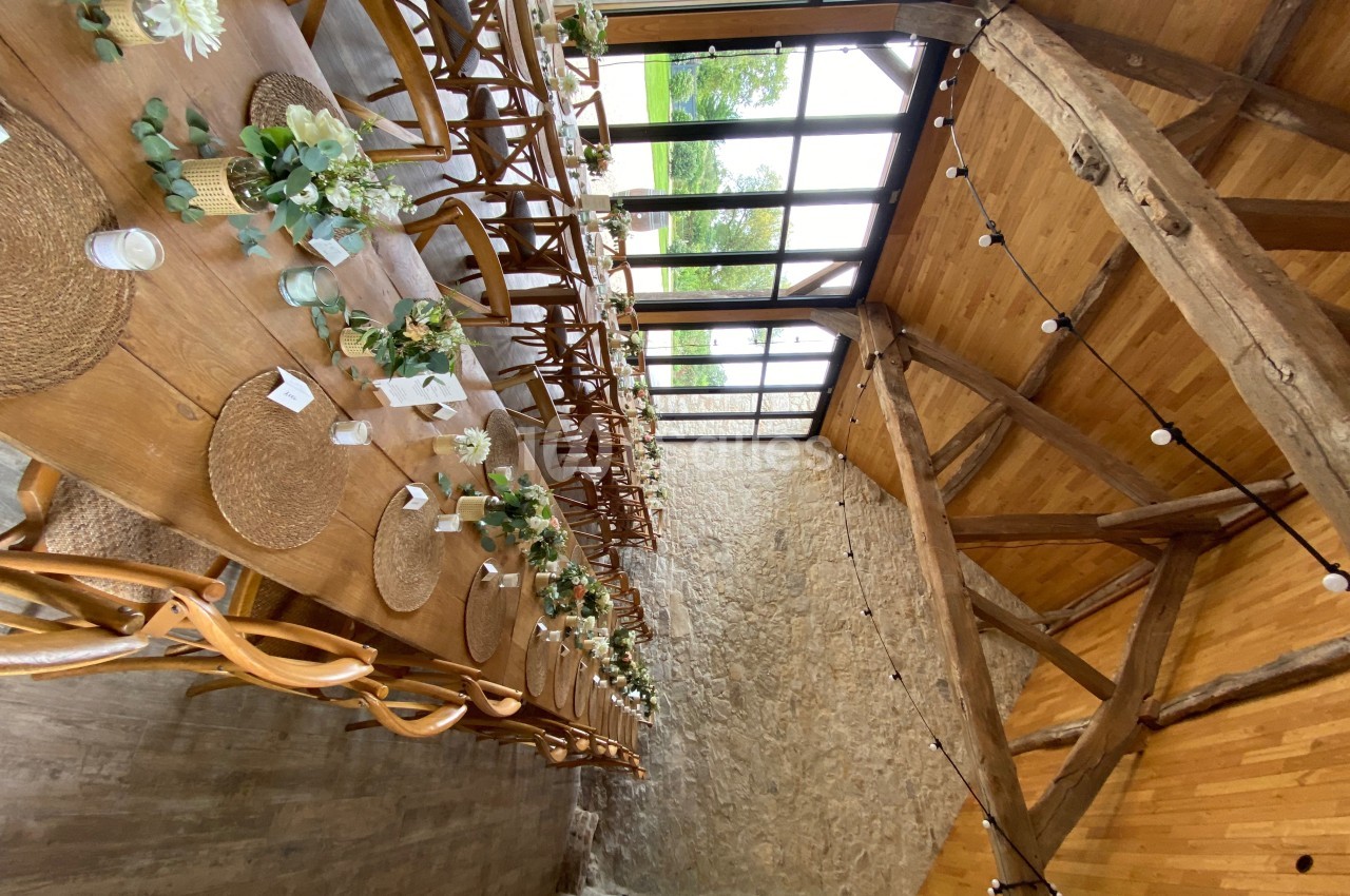 Grande table en bois décorée de fleurs et de sets en paille dans une salle avec poutres apparentes et grandes baies vitrées.