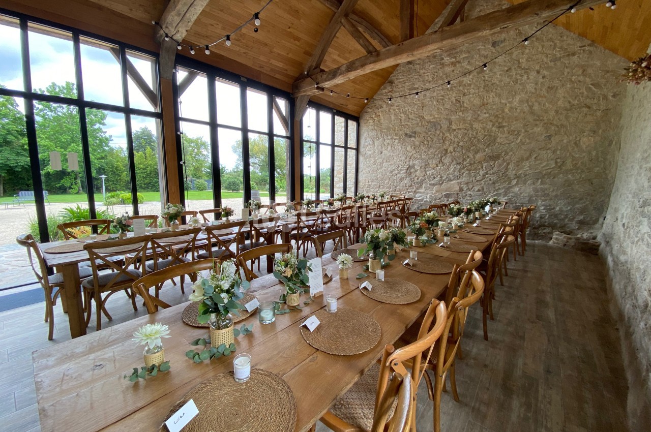 Salle de réception avec tables en bois décorées de fleurs, sous une charpente apparente et près de grandes baies vitrées.