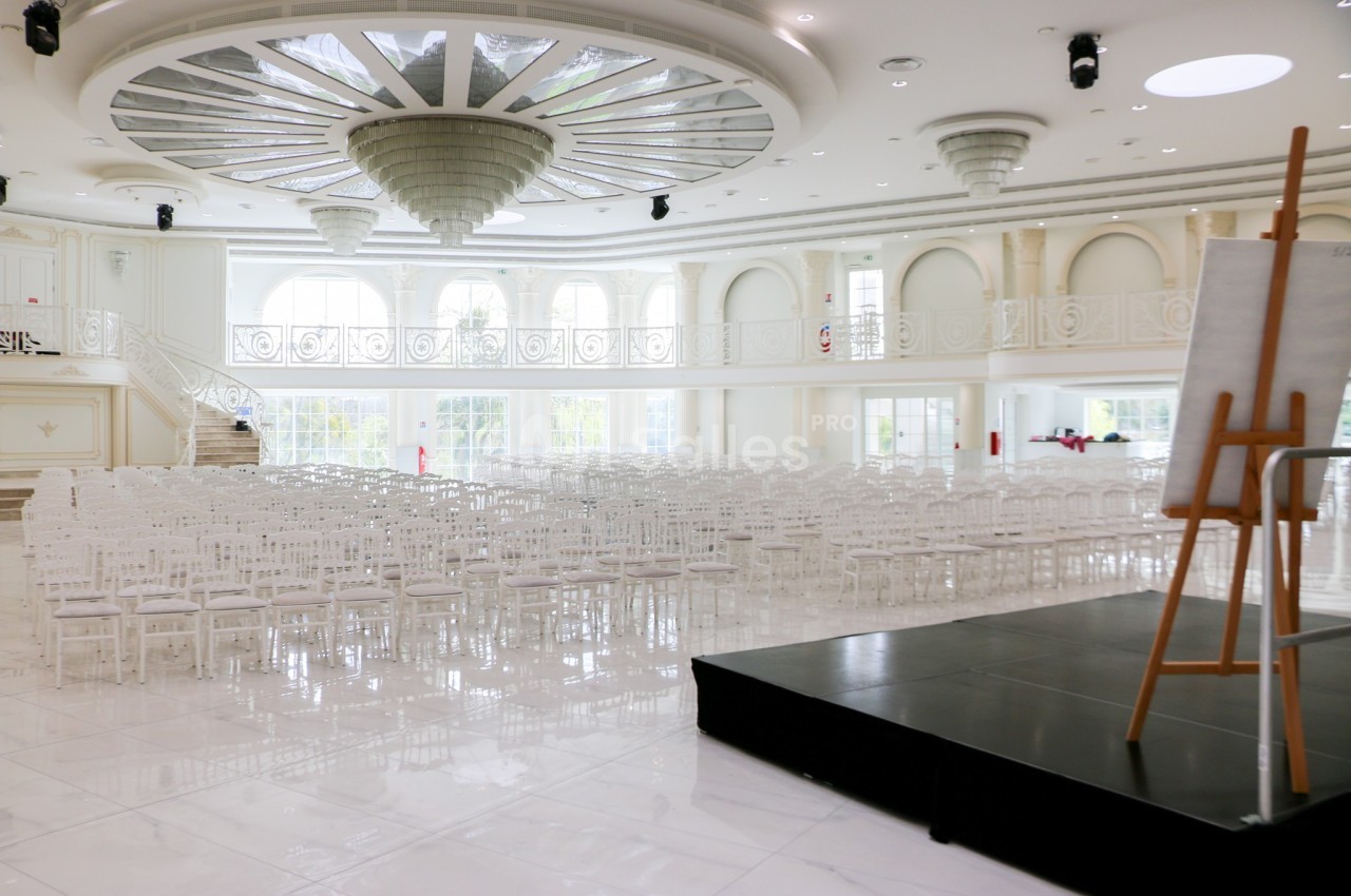 Salle de réception lumineuse avec des chaises blanches alignées, un podium noir et un chevalet à droite.