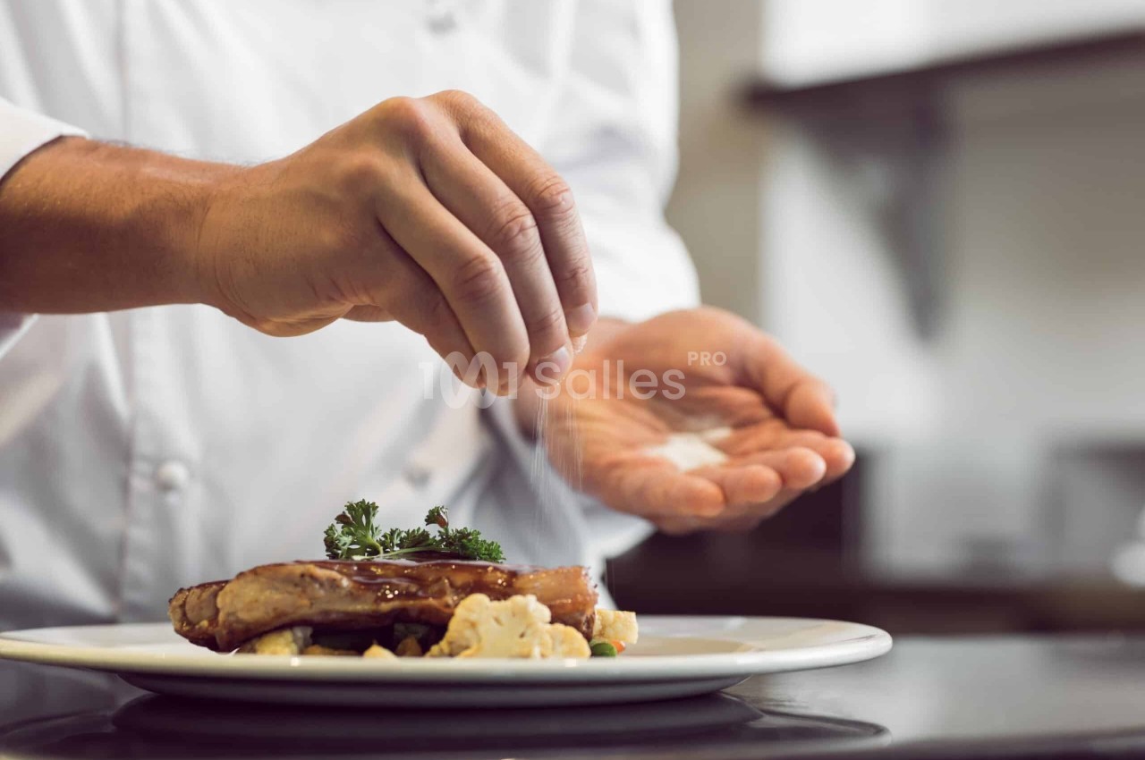 Un chef saupoudre du sel sur un plat garni de viande, légumes et herbes dans une cuisine professionnelle.