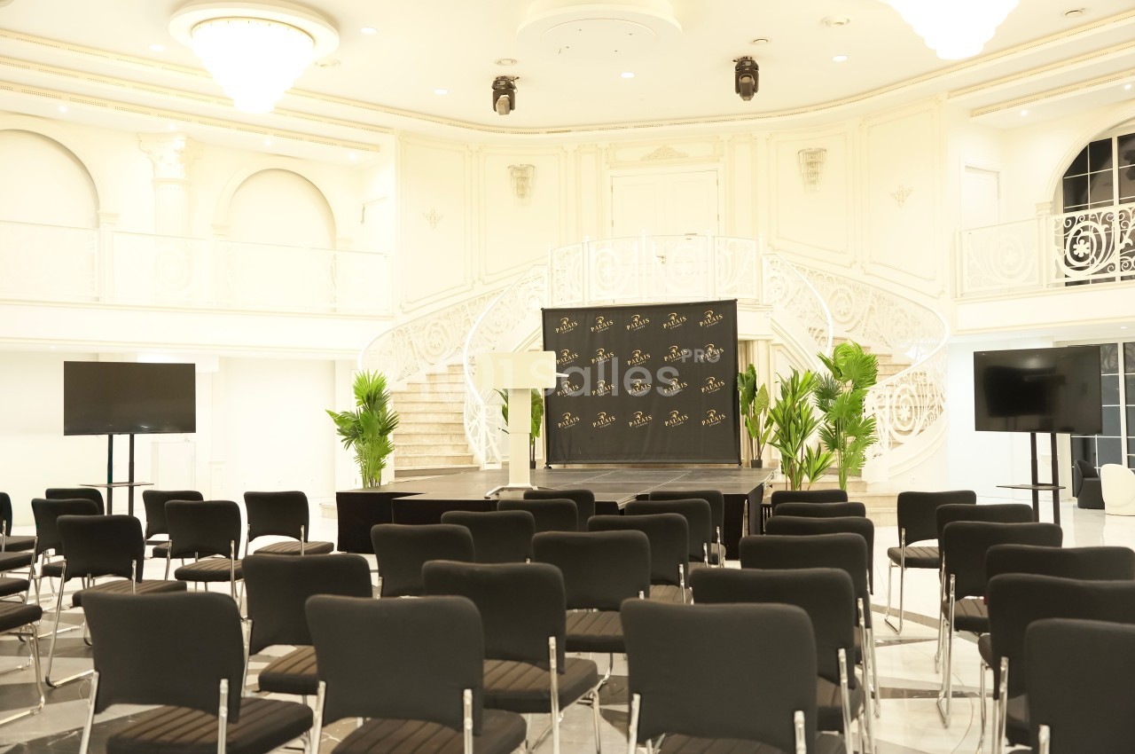 Salle de conférence élégante avec chaises noires alignées, scène centrale, plantes vertes et escalier en arrière-plan.