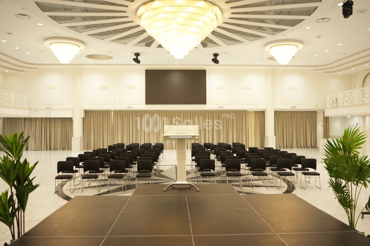 Salle de conférence lumineuse avec un podium, des rangées de chaises noires et un grand écran en arrière-plan.