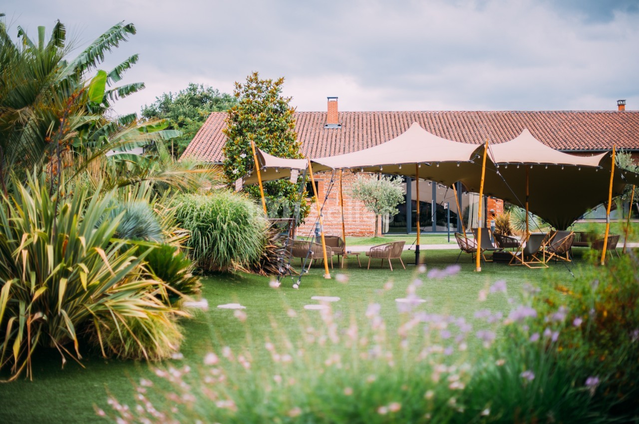 Jardin paysagé avec tonnelles aménagées, chaises, maison en brique aux tuiles terracotta et pelouse fleurie.
