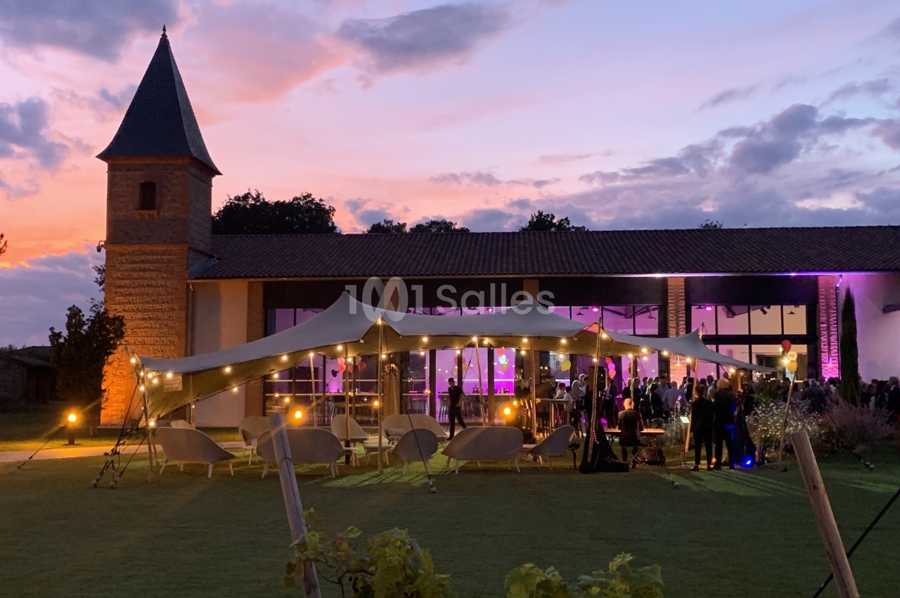 Salle de réception en terrasse avec voile tendu illuminé, convives debout et assis, bâtiment en briques et tour, crépuscule.