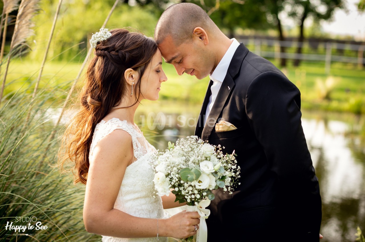 Mariés en robe blanche et costume noir avec bouquet blanc posent dans un jardin verdoyant bordé d'eau.