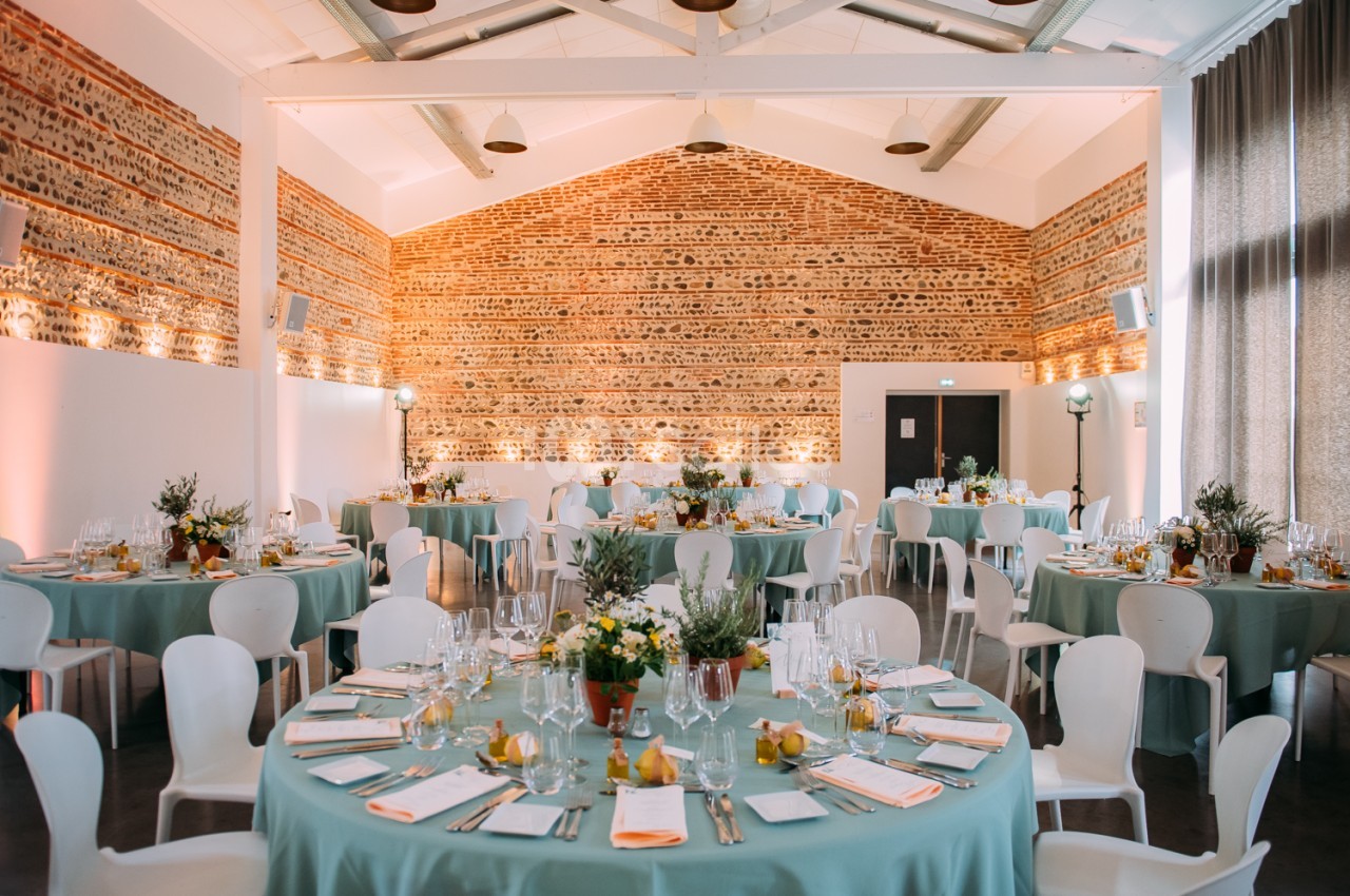 Salle intérieure dressée avec tables rondes en nappe turquoise, chaises blanches, murs en brique rouge et plafond blanc - Mas Tolosa.