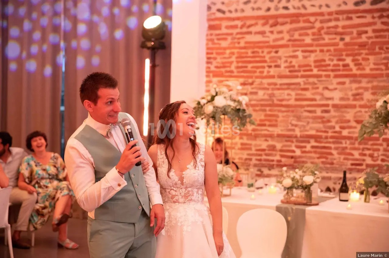 Salle intérieure avec mur de briques rouges, éclairage ambiant rose-violet, tables dressées et couple au centre.