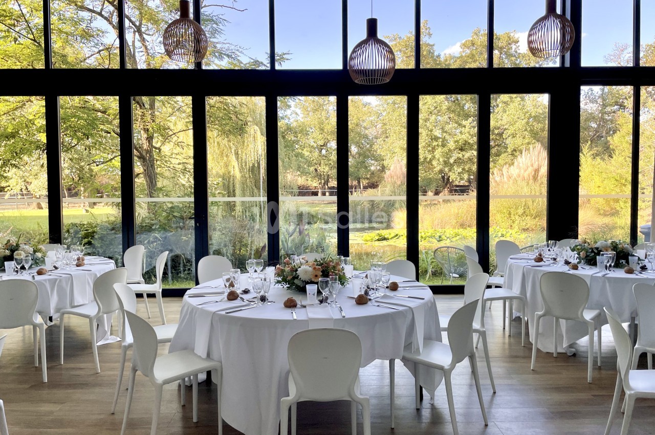 Salle de réception avec tables rondes dressées en blanc, chaises blanches et baies vitrées donnant sur parc arboré.