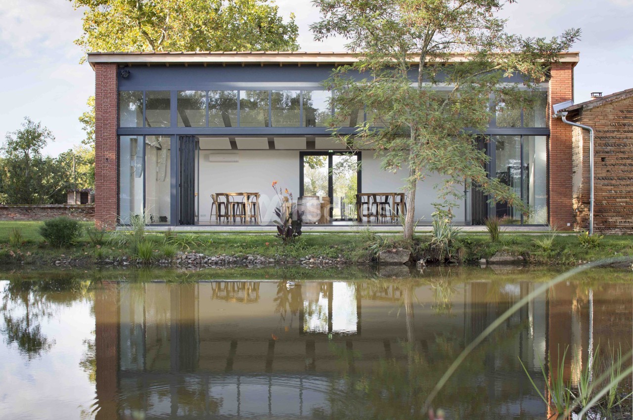 Bâtiment contemporain en brique avec grandes baies vitrées surplombant un plan d'eau, intérieur aménagé de tables et chaises.