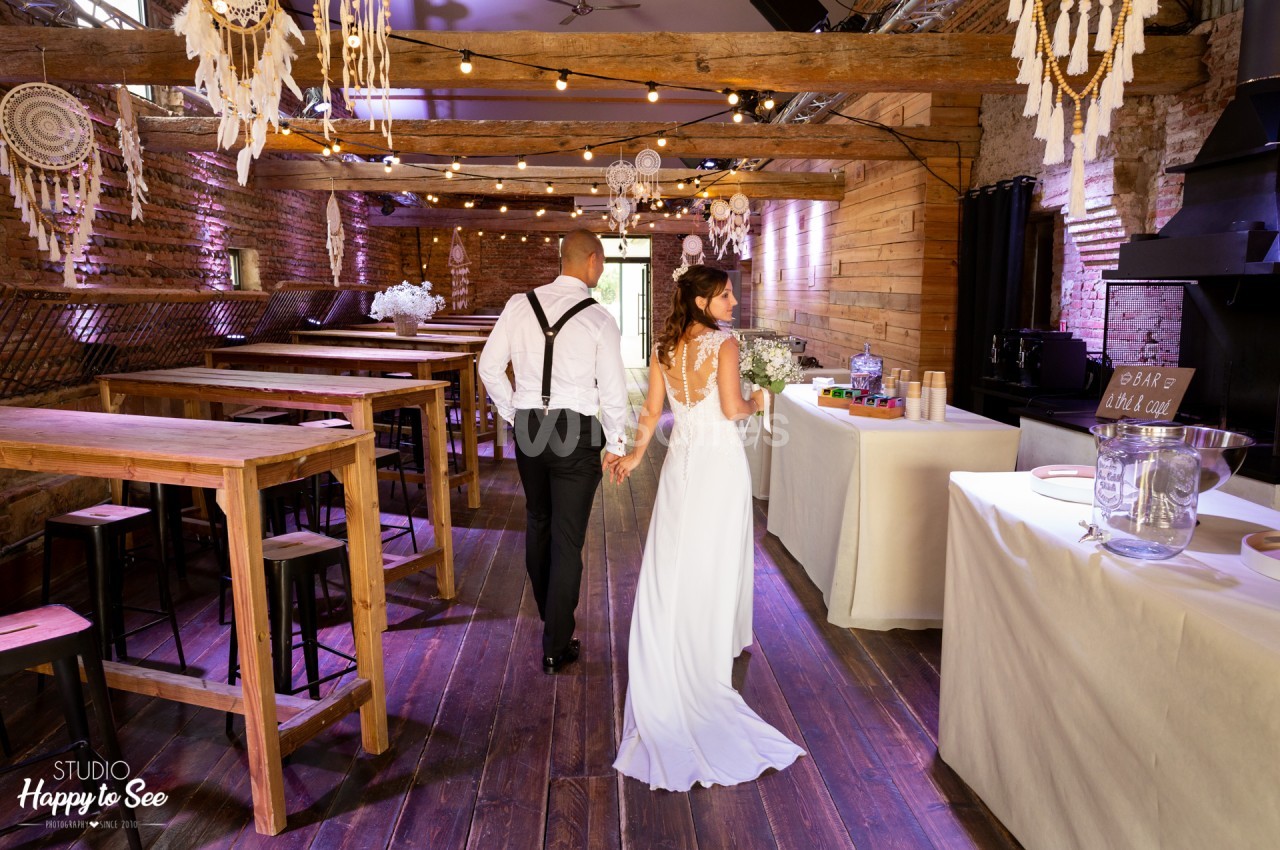 Salle en pierre et bois avec poutres apparentes, guirlandes lumineuses et dreamcatchers, couple en tenue de mariage circulant entre tables dressées.