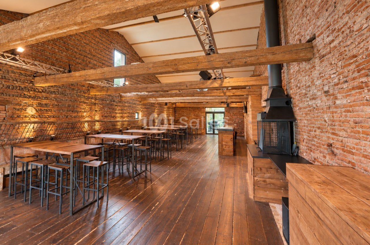 Salle intérieure en briques avec poutres bois, tables hautes et chaises, poêle noir, éclairage spots.