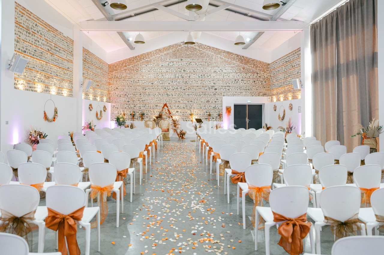 Salle de cérémonie intérieur avec chaises blanches décorées de nœuds oranges, murs en brique, allée fleurie.