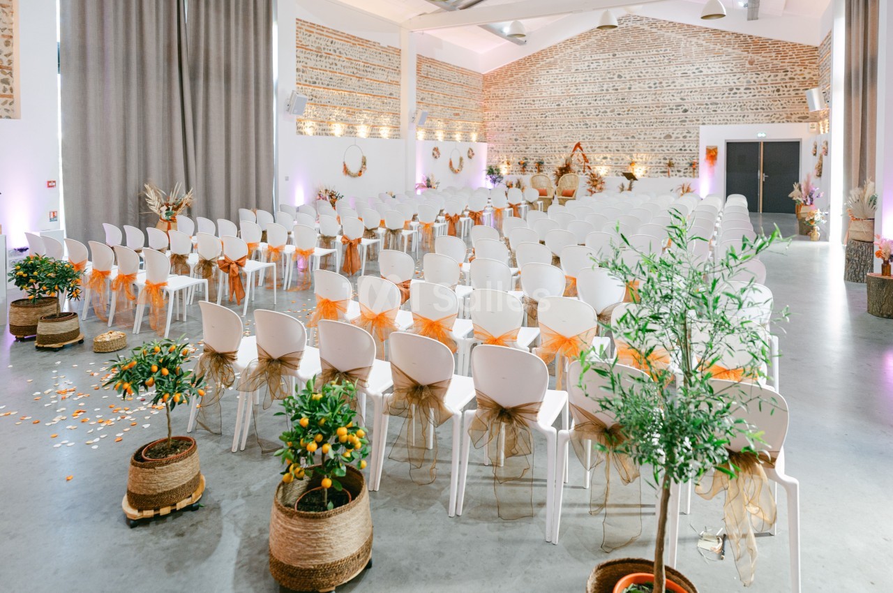Salle intérieure aménagée en cérémonie avec chaises blanches décorées de nœuds oranges, plantes en pots et murs en briques apparentes.