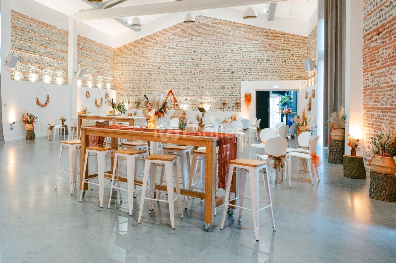 Salle à haut plafond avec murs en briques apparentes, tables hautes en bois, chaises blanches et décoration florale automnale.