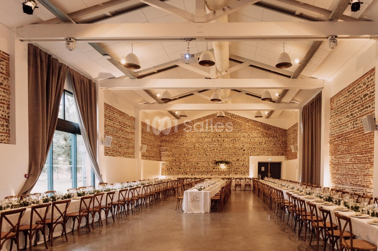 Salle intérieure avec murs en briques, tables dressées en U et chaises en bois, plafond apparent aux poutres peintes.