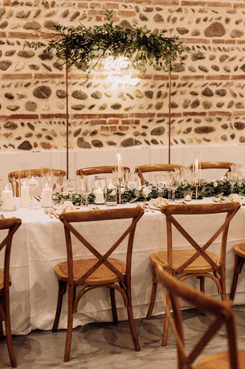 Salle dressée avec tables blanches, chaises en bois, mur en pierre et décor végétal suspendu.