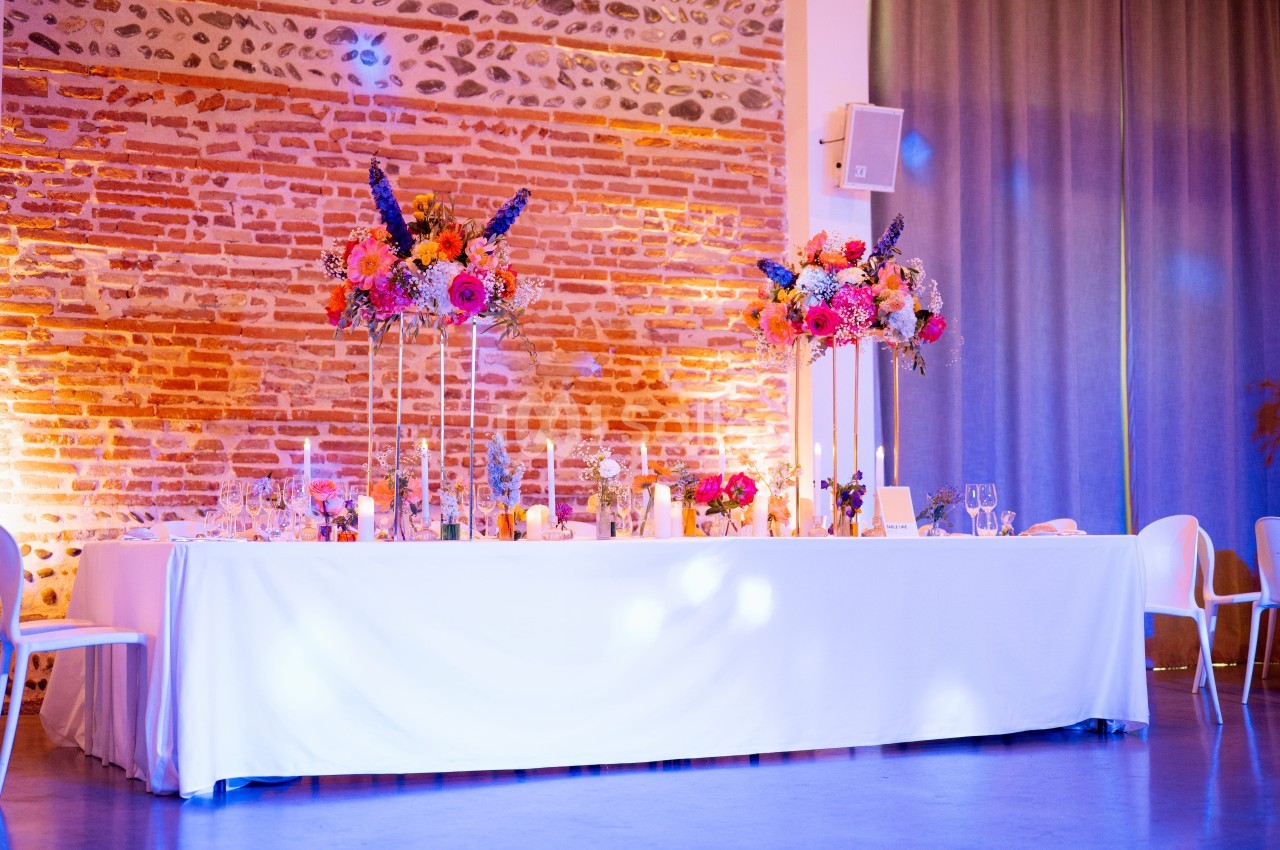 Table dressée avec nappe blanche, compositions florales colorées sur pieds hauts, mur de briques illuminé rose et or, ambiance événementielle.