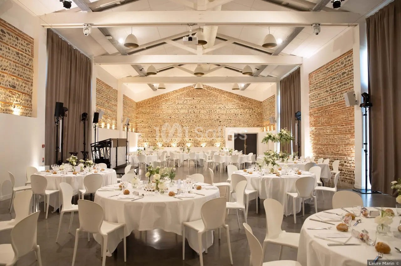 Salle intérieur avec murs briques apparentes, tables rondes dressées nappes blanches et chaises blanches, décoration florale épurée.