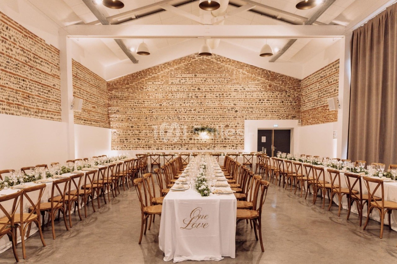 Salle intérieure avec murs en briques apparentes, plafond blanc à poutres, tables dressées en U avec chaises en bois et fleurs blanches.