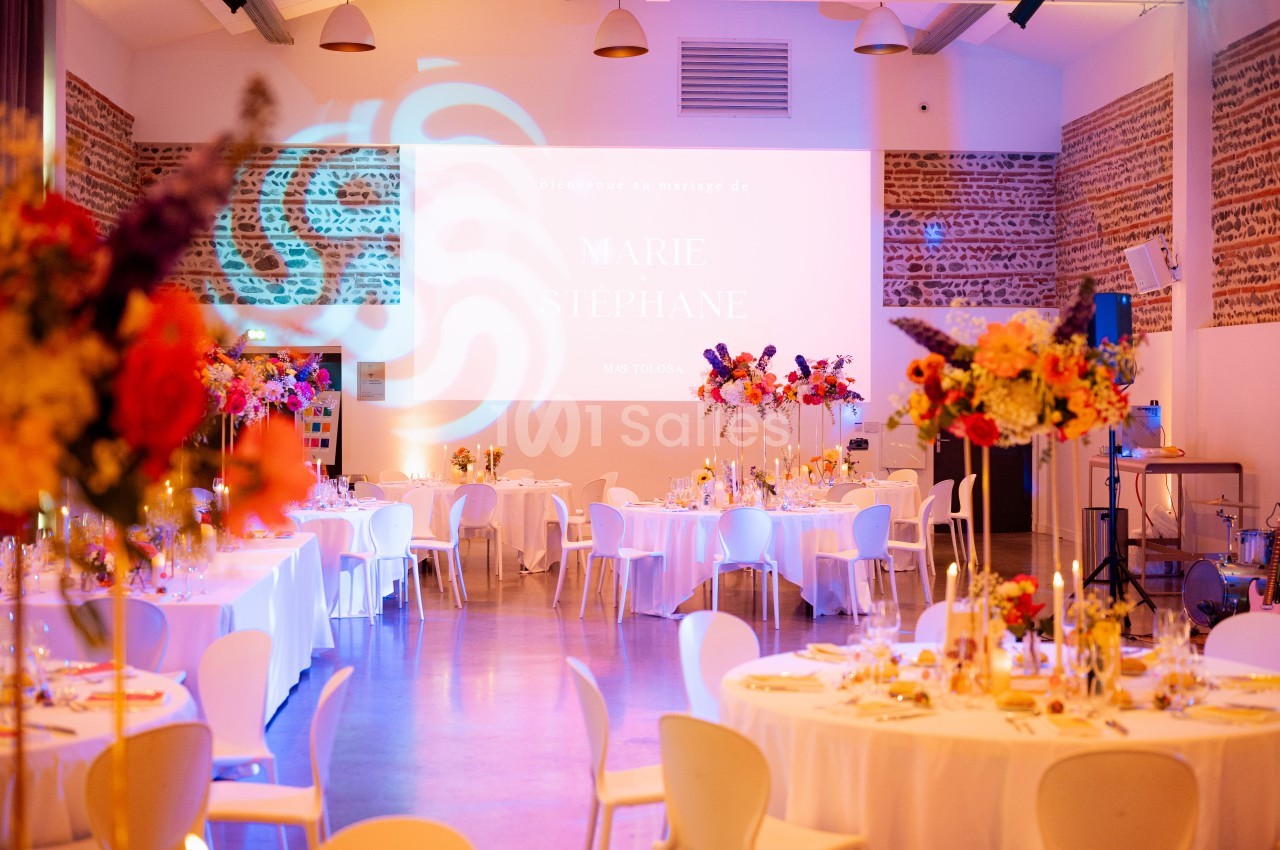 Salle aménagée avec tables rondes dressées, chaises blanches, éclairage rose-violet et bouquets de fleurs colorées.