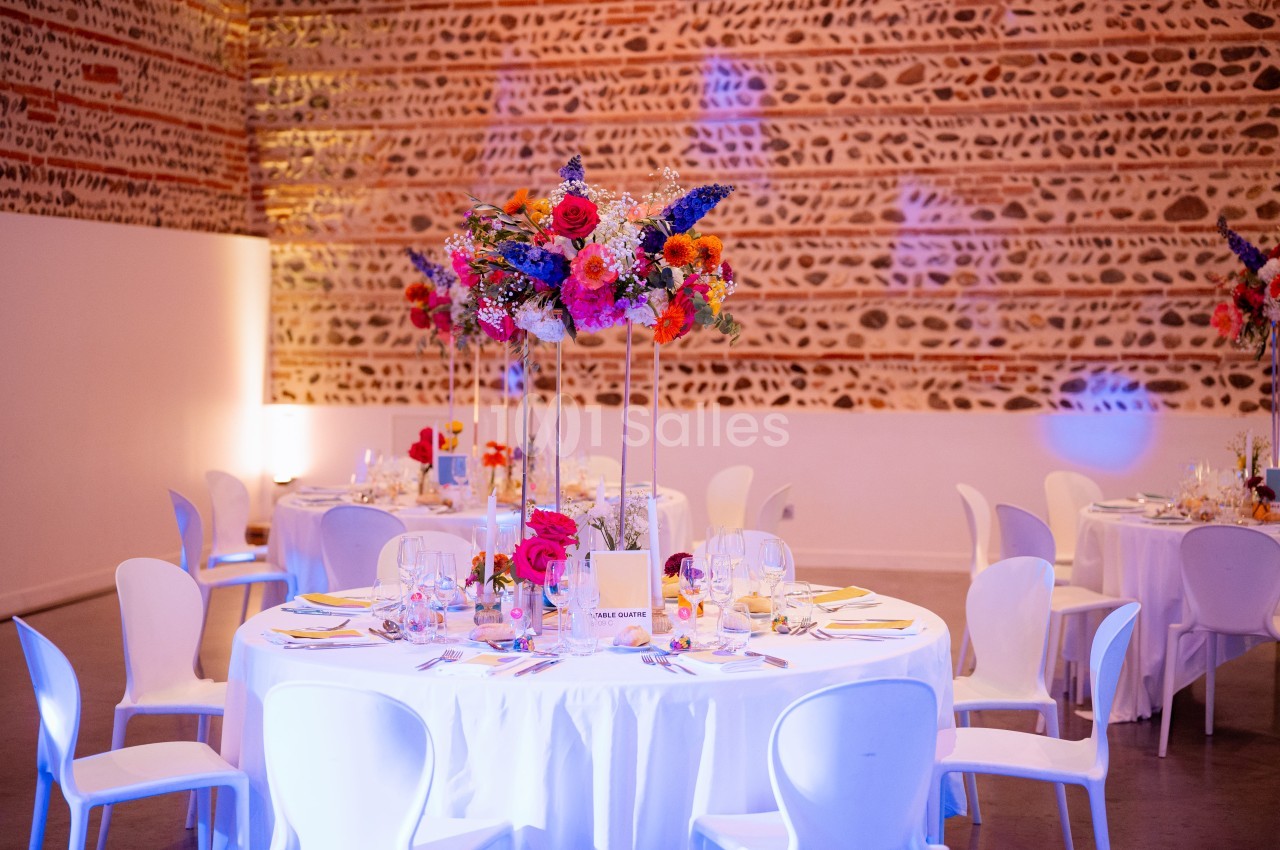 Salle au plafond ajouré en briques avec tables rondes dressées, chaises blanches et compositions florales colorées sur hauts vases.