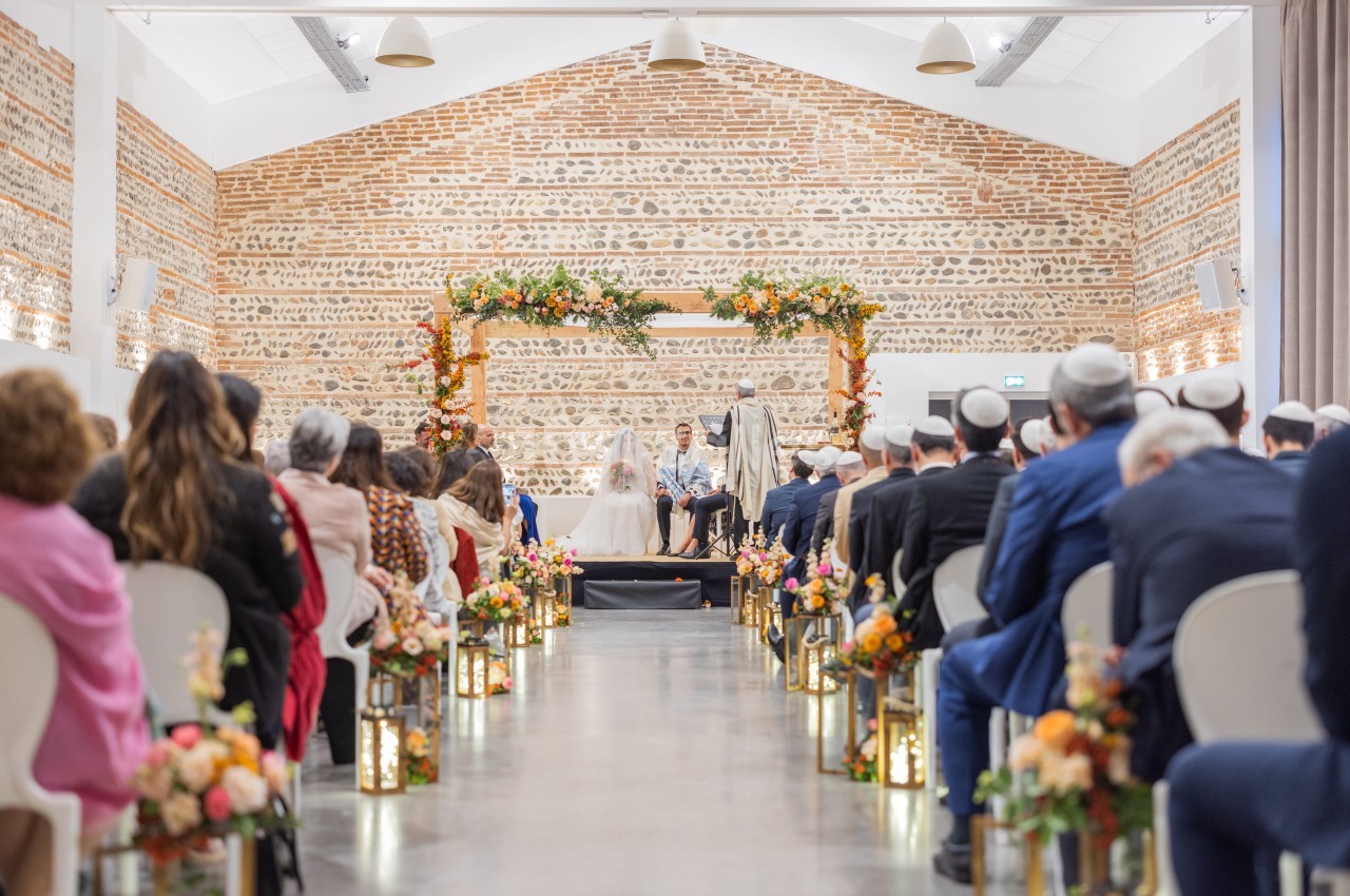 Cérémonie de mariage dans une salle aux murs en pierre brute, convives assis face à l'autel fleuri.