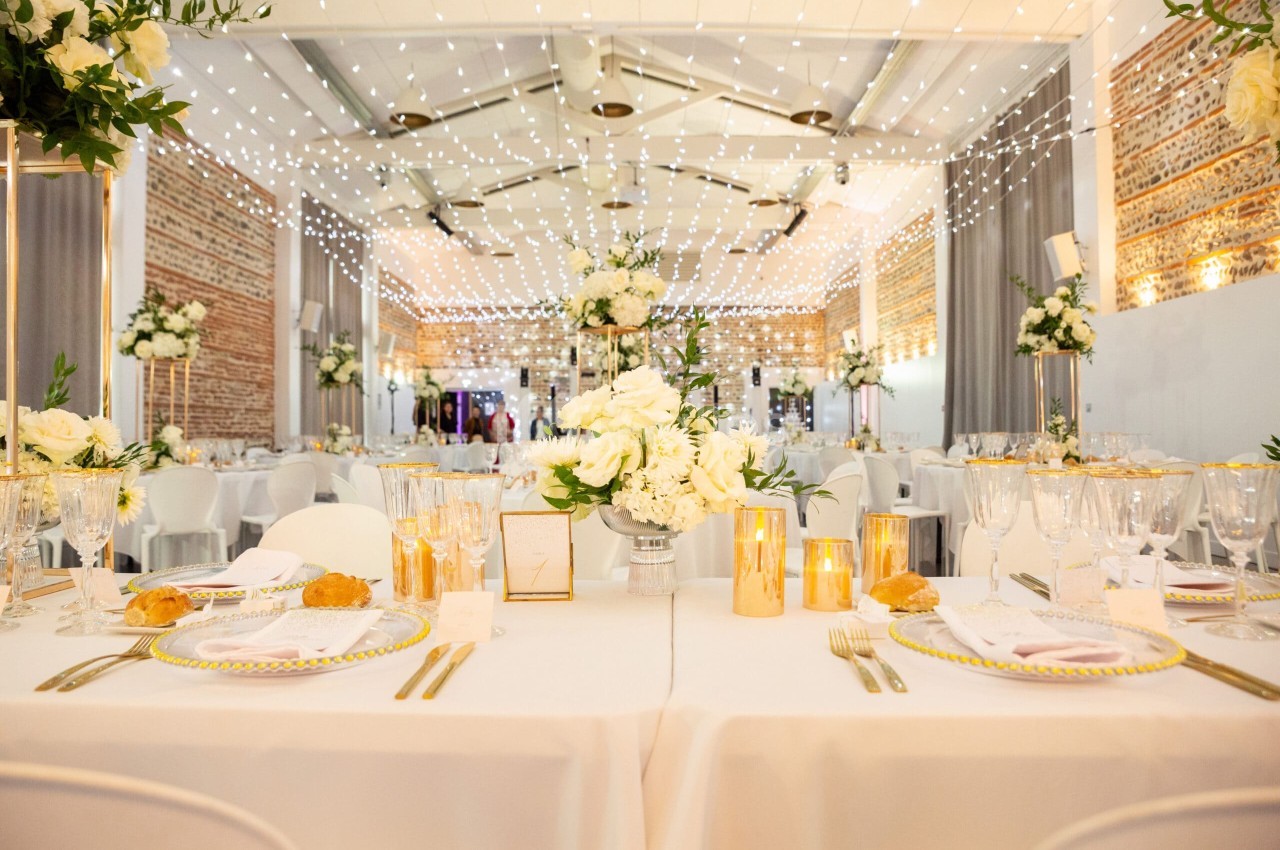 Salle dressée avec tables blanches, décoration florale blanche et or, guirlandes lumineuses au plafond, murs briques apparentes.