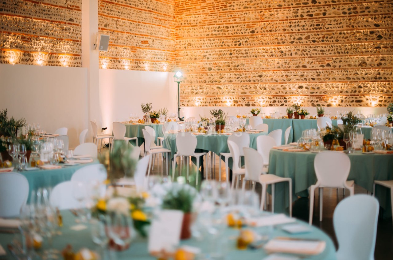 Salle dressée pour banquet avec tables rondes aux nappes vert d'eau, chaises blanches et décoration florale - murs en pierre brute éclairés.