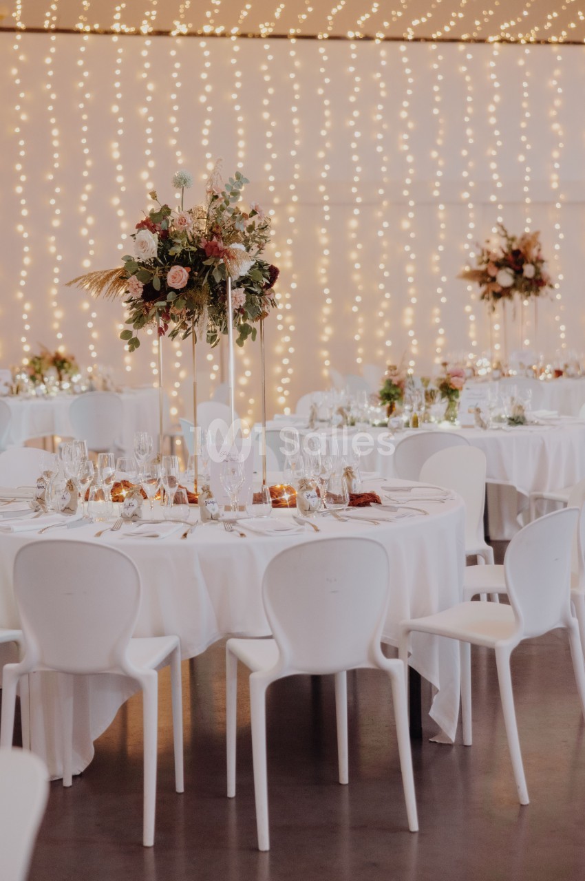 Salle de réception dressée avec tables rondes blanches, chaises blanches, fond lumineux de guirlandes de perles et compositions florales élevées.
