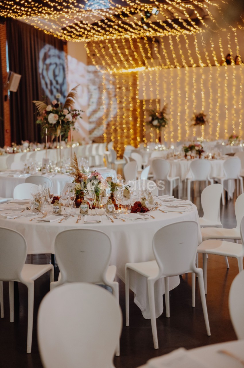 Salle de réception dressée avec tables rondes blanches, chaises blanches, lustres dorés au plafond et bouquets de fleurs.