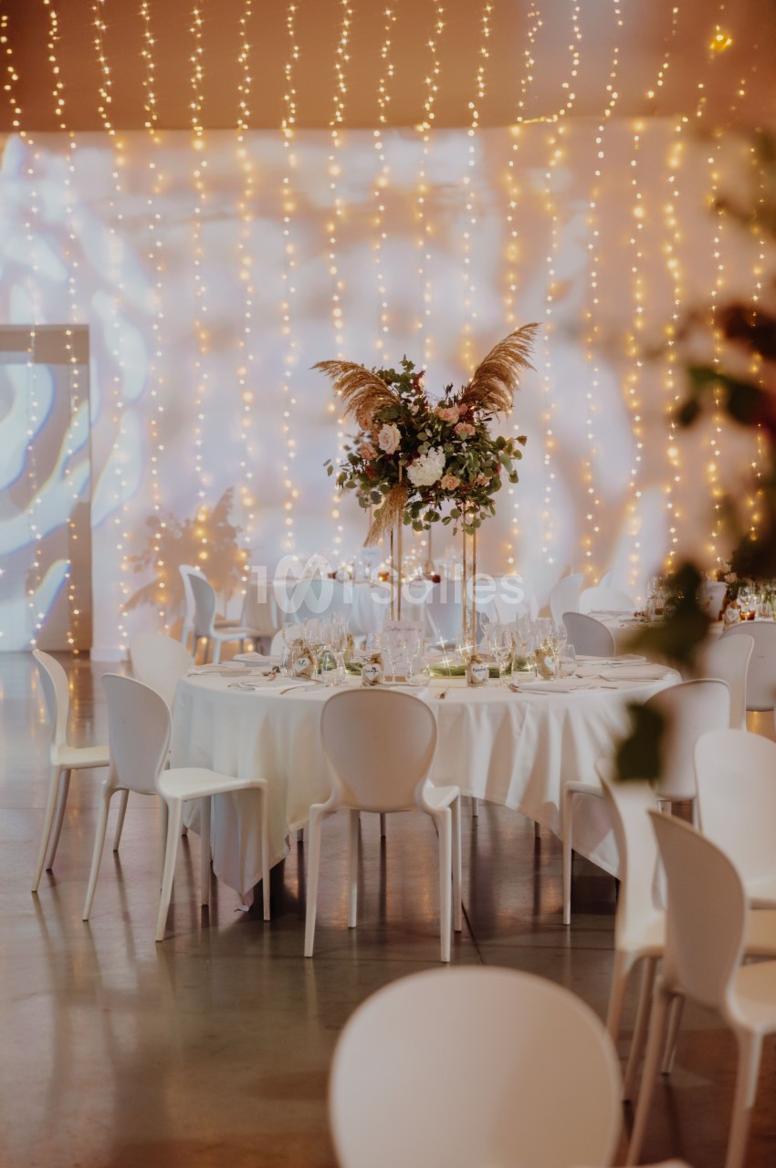 Salle de réception intérieure dressée avec tables rondes blanches, chaises blanches, tentures lumineuses dorées et arrangement floral central aux teintes roses et cuivrées.