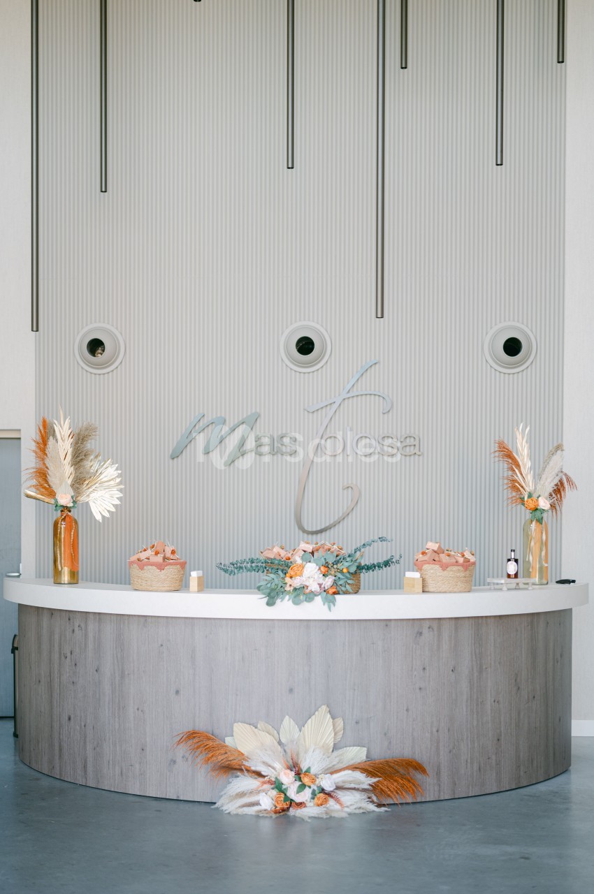 Comptoir de réception courbe en bois clair, orné de bouquets de fleurs orange et blanches, de vases en verre et de bougies.