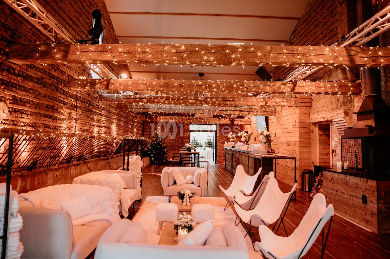 Salle intérieure en briques avec poutres apparentes, guirlandes lumineuses, fauteuils et canapés blanc cassé, bar au fond.