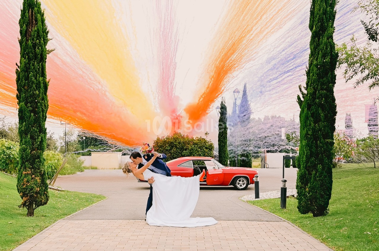 Couple en tenue de mariage dans une allée bordée de cyprès, voiture vintage rouge en arrière-plan.