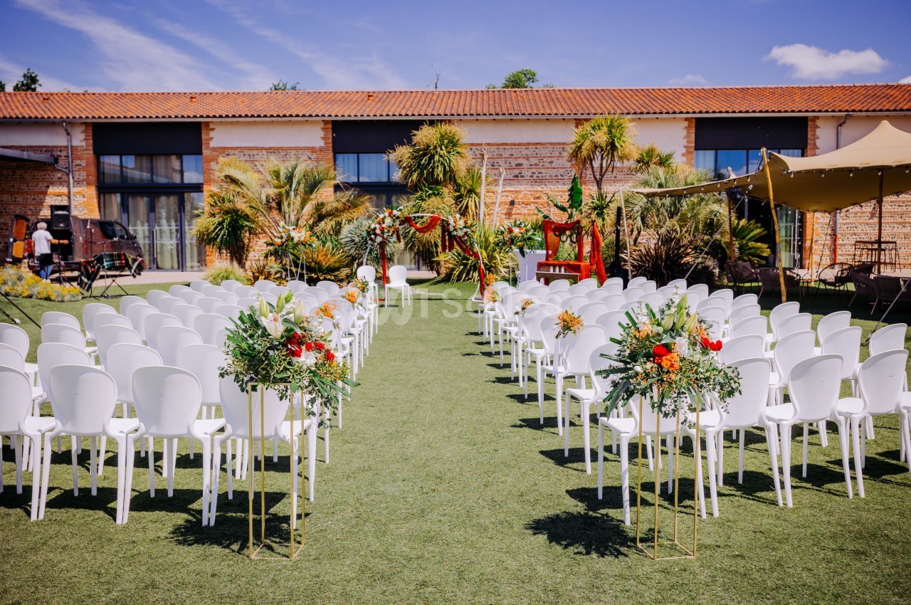 Cérémonie laïque en plein air - chaises blanches alignées face à un autel fleuri devant bâtisse en brique.