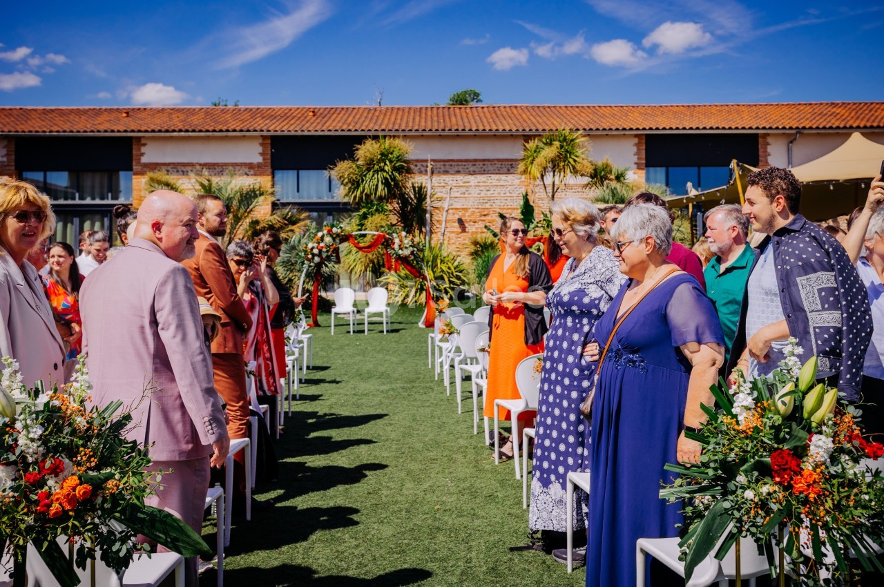 Cérémonie en extérieur dans le jardin du Mas avec chaises blanches alignées, fleurs oranges, convives debout et bâtiment en pierre en arrière-plan.