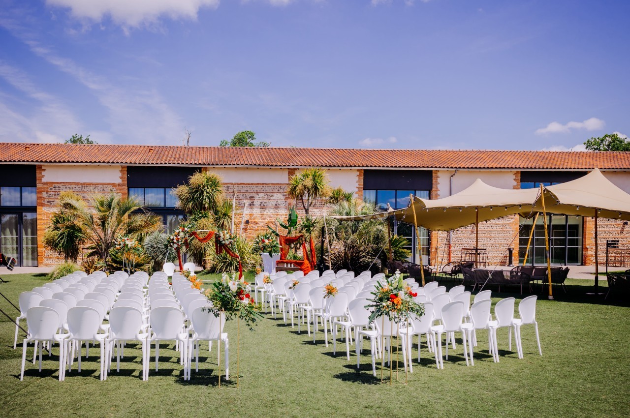 Cérémonie en plein air avec chaises blanches, allée fleurie et bâtiment en brique sous ciel bleu.