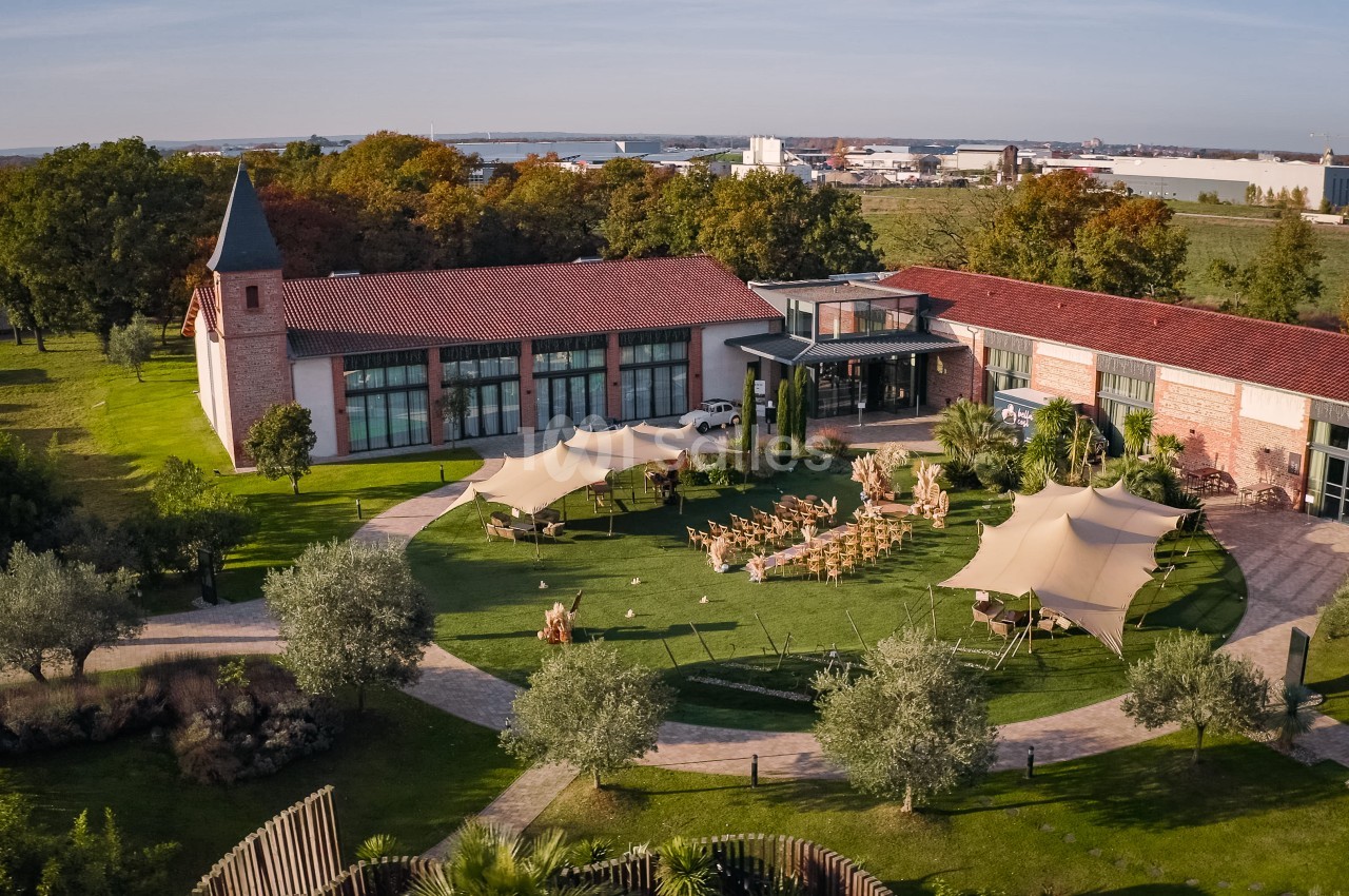 Vue aérienne du domaine avec bâtiment principal en brique, jardin aménagé, chaises disposées pour réception et voiles blanches.