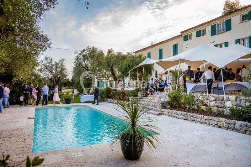 Jardin avec piscine, tente blanche et invités rassemblés près d'une maison en pierre sous un ciel partiellement nuageux.