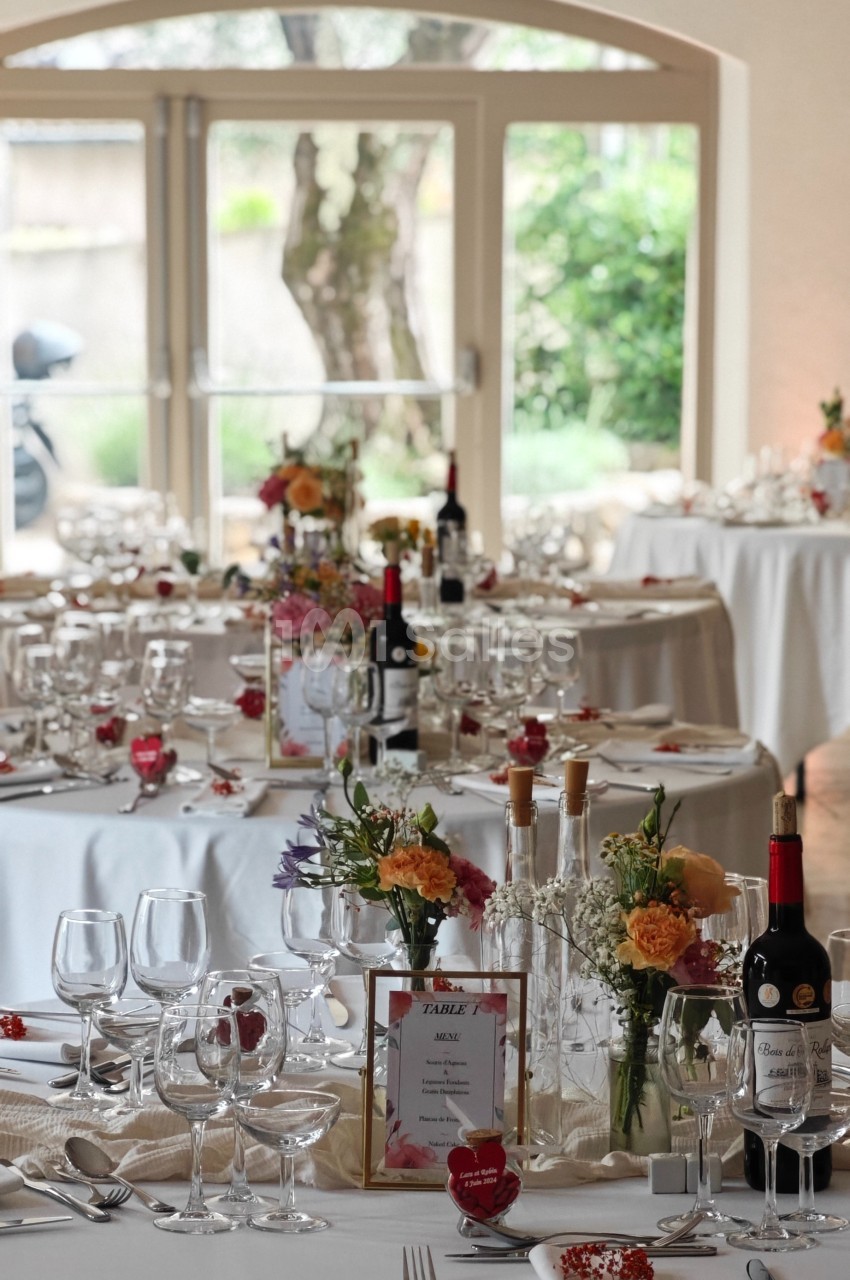 Tables élégamment dressées avec nappes blanches, fleurs colorées, verres et bouteilles de vin dans une salle lumineuse.