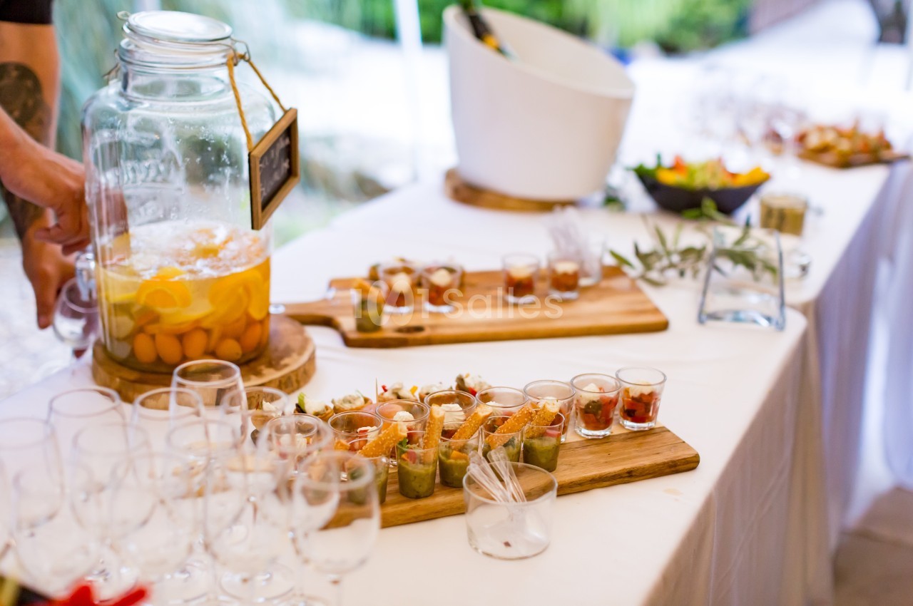 Buffet avec verrines, carafe de boisson aux fruits et seaux à glace, disposés sur une table blanche en extérieur.