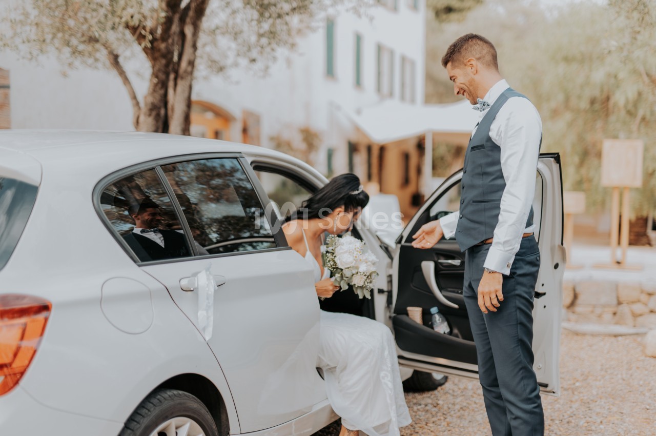 Une femme en robe blanche sort d'une voiture décorée, aidée par un homme en costume, dans un cadre extérieur.