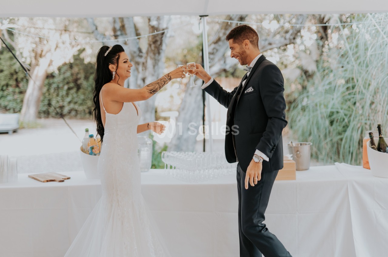 Un couple en tenue de mariage trinque sous une tente blanche, avec une table dressée en arrière-plan.