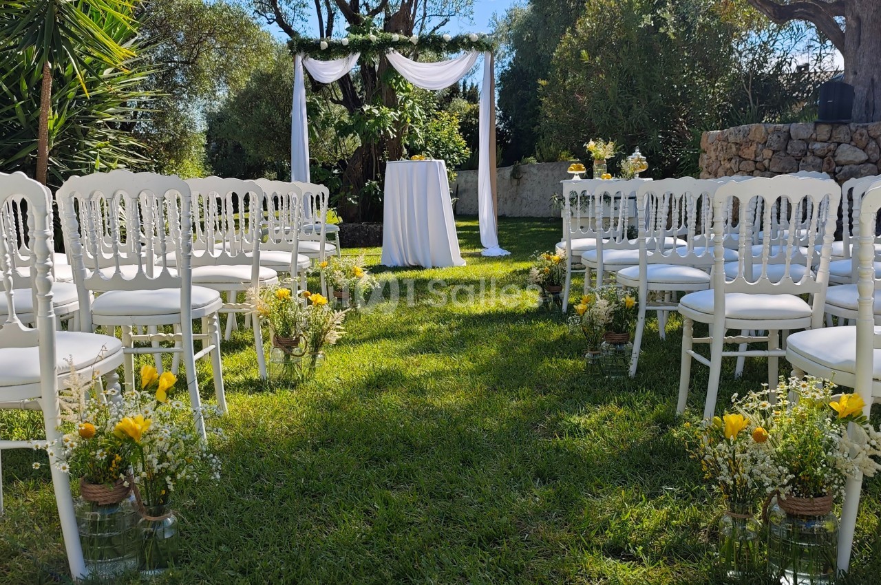 Chaises blanches alignées face à une arche décorée de tissu blanc et fleurs, installées dans un jardin ensoleillé.