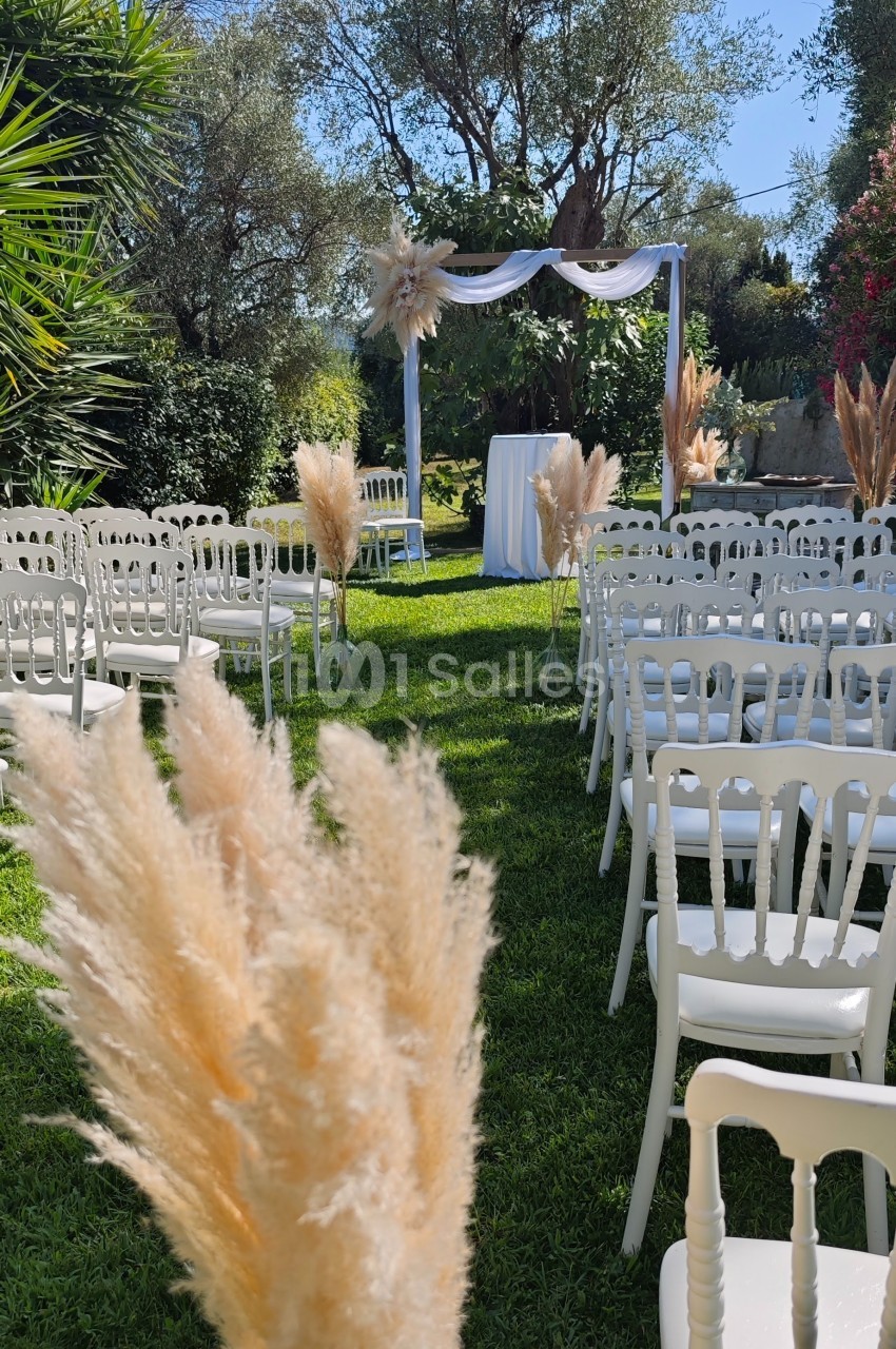 Chaises blanches alignées sur une pelouse entourée de végétation, avec une arche décorée pour une cérémonie en plein air.
